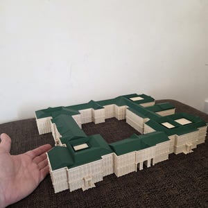May include: A detailed architectural model of a building with a green roof and off-white walls. The model is a rectangular shape with multiple sections and appears to be a miniature replica of a large structure. The model is on a brown surface.