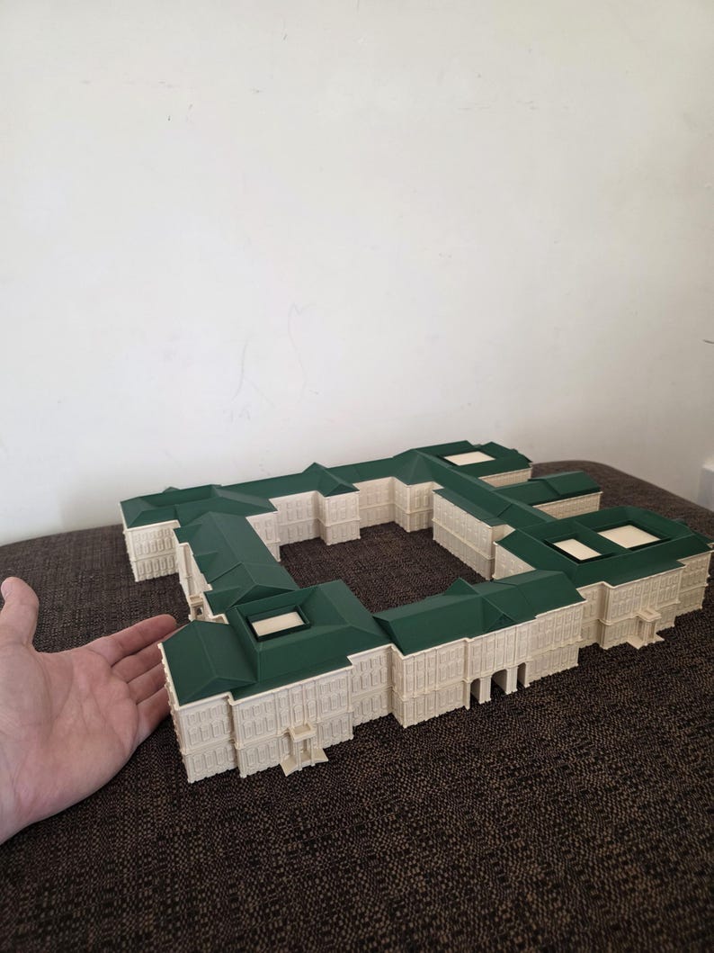 May include: A detailed architectural model of a building with a green roof and off-white walls. The model is a rectangular shape with multiple sections and appears to be a miniature replica of a large structure. The model is on a brown surface.