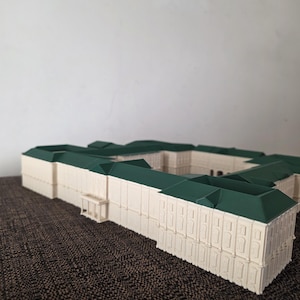May include: A detailed miniature building model with a cream-coloured facade and a dark green roof. The model has a rectangular shape with multiple sections and architectural details. It is placed on a textured brown surface.