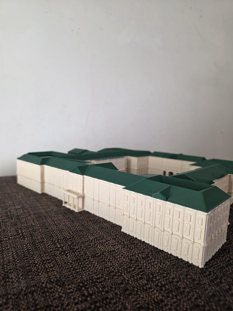 May include: A detailed miniature building model with a cream-coloured facade and a dark green roof. The model has a rectangular shape with multiple sections and architectural details. It is placed on a textured brown surface.
