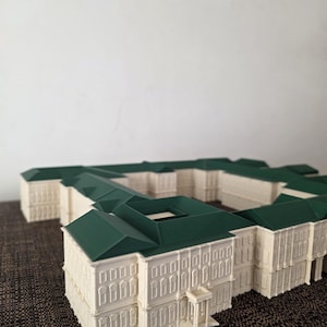 May include: A detailed architectural model of a building with a cream-coloured facade and a dark green roof. The model features multiple rectangular sections, with rows of windows and decorative elements. The building has a symmetrical design and a small entrance.