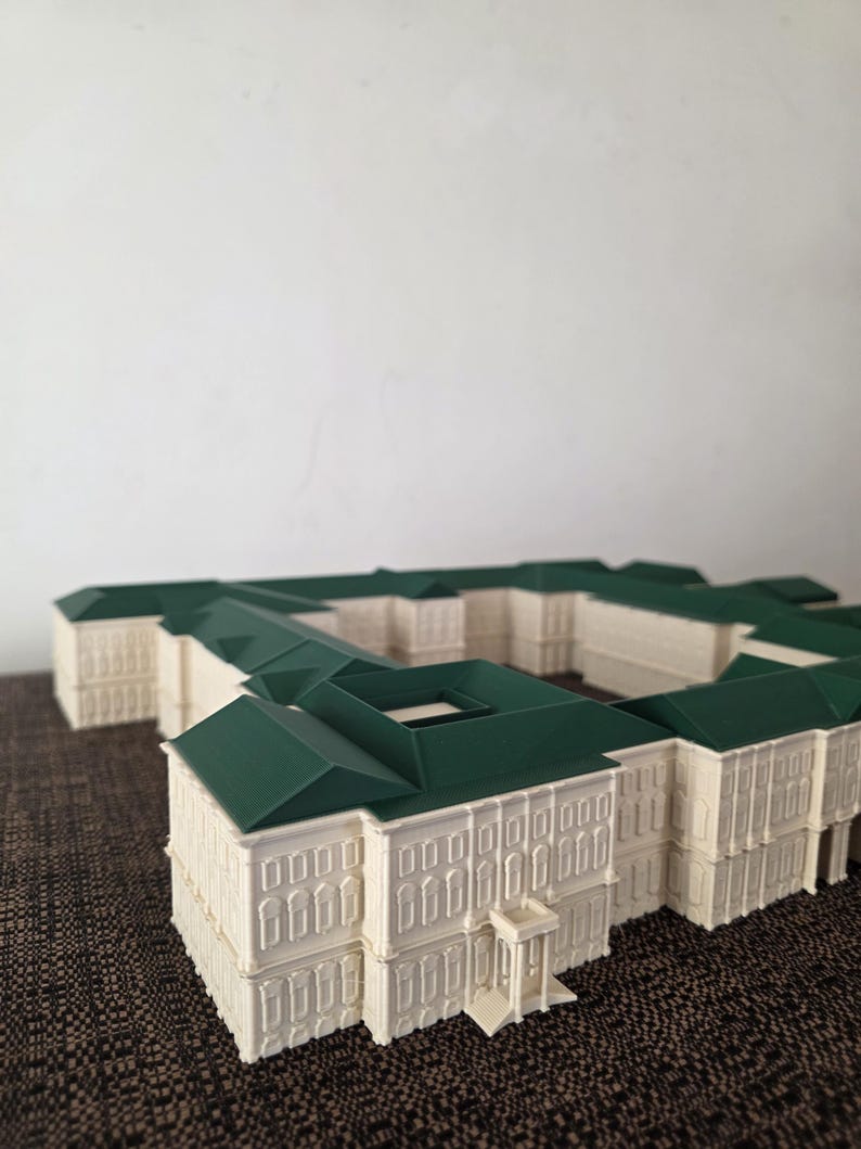 May include: A detailed architectural model of a building with a cream-coloured facade and a dark green roof. The model features multiple rectangular sections, with rows of windows and decorative elements. The building has a symmetrical design and a small entrance.