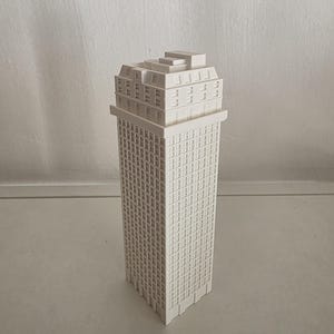 Könnte beinhalten: Ein weißes, 3D-gedrucktes Architekturmodell eines hohen Wolkenkratzers mit rechteckiger Grundfläche, einer Fassade mit zahlreichen quadratischen Fenstern und einer gestuften Dachstruktur mit kleineren Fenstern.