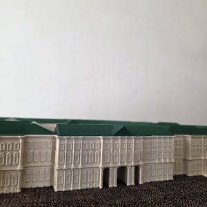 May include: A miniature model of a building with a white facade and a green roof. The building has multiple windows and arched entryways. The model is set against a neutral background.