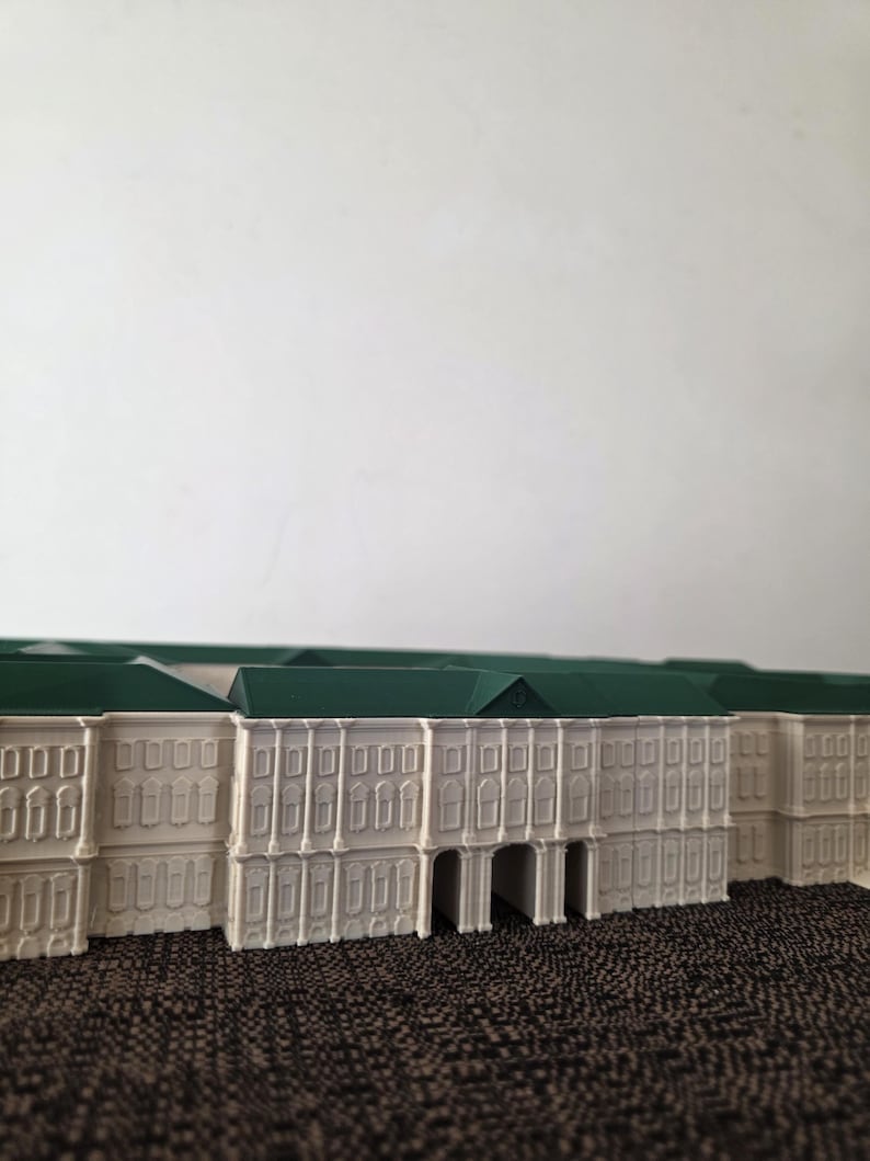 May include: A miniature model of a building with a white facade and a green roof. The building has multiple windows and arched entryways. The model is set against a neutral background.