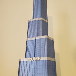 Puede incluir: Maqueta de un rascacielos alto con fachada azul y la palabra "TRUMP" en letras blancas. El edificio tiene una base rectangular y una aguja en la parte superior. Las ventanas son pequeñas y espaciadas uniformemente.