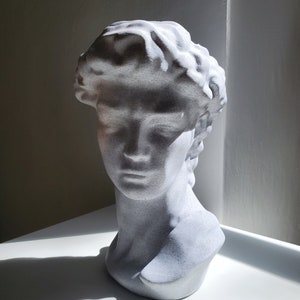 May include: White plaster bust sculpture of a person's head and shoulders. The sculpture has a textured surface and is in a minimalist style.