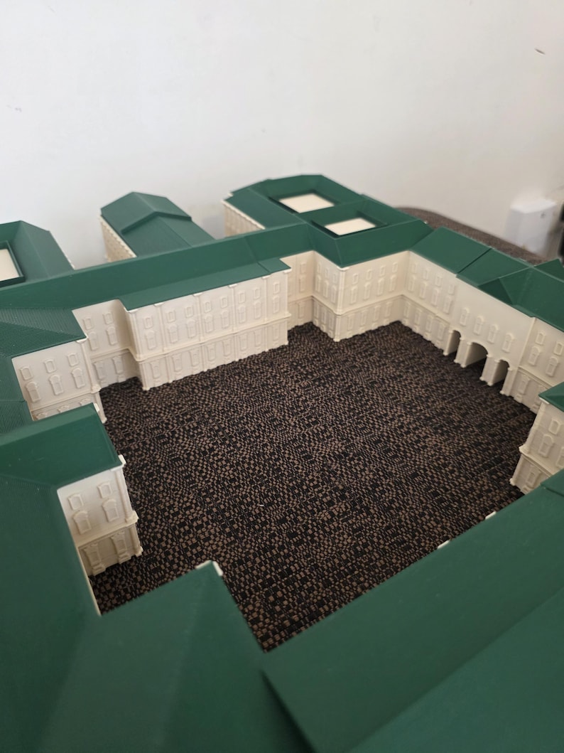 May include: A detailed miniature model of a building complex. The model features white walls, green roofs, and a central courtyard. The building has multiple windows and a detailed design.