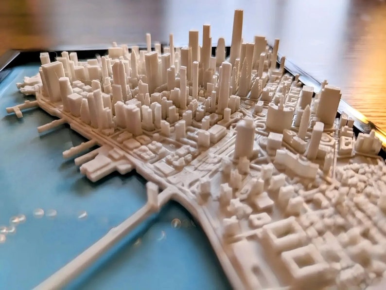 Op de afbeelding: Een wit 3D-geprint model van een stadsbeeld met gebouwen en straten. Het model staat op een blauwe basis en is omgeven door een zwart frame.