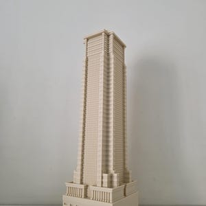 Può includere: Modello architettonico beige di un grattacielo. L'edificio presenta una base rettangolare con colonne, un'alta torre a più piani con una griglia di finestre e un tetto piatto. Il modello è realizzato in materiale chiaro.