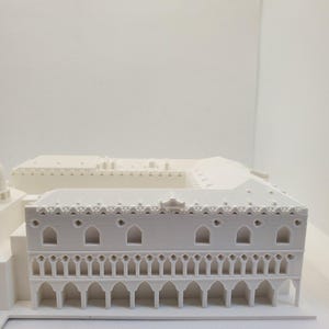 May include: A white 3D printed model of a building with intricate details. The building has a flat roof with a decorative border and a series of arches along the bottom.