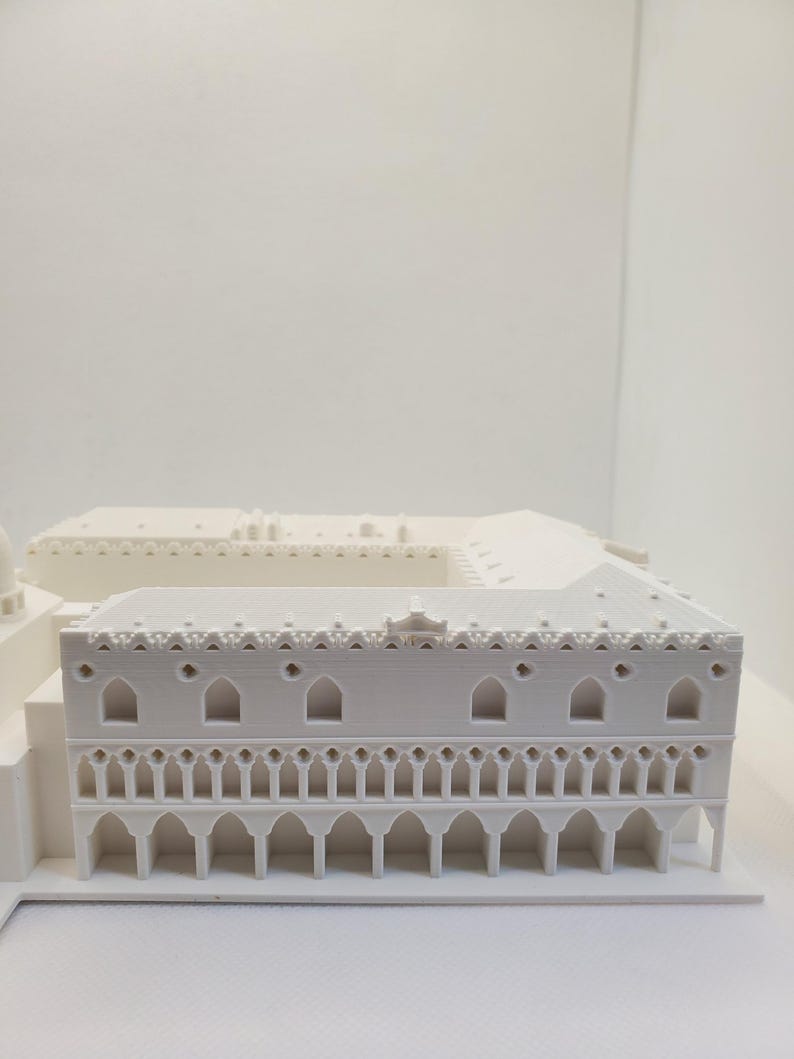 May include: A white 3D printed model of a building with intricate details. The building has a flat roof with a decorative border and a series of arches along the bottom.
