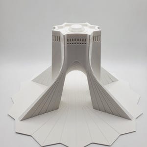 Op de afbeelding: Een wit 3D-geprint model van de Azadi-toren in Teheran, Iran. De toren is een stervormige structuur met een koepelvormig dak en een grote boog. Het model staat op een stervormige basis.