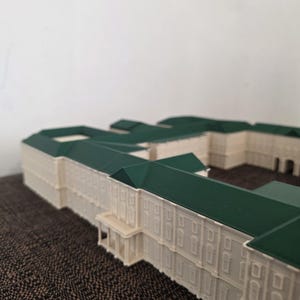 May include: A detailed architectural model of a building with a cream facade and a dark green roof. The model features intricate details, including windows and a small porch. The building is set against a plain background.