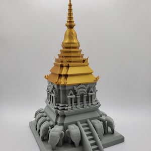May include: A 3D printed model of a golden pagoda with a gray base. The base is decorated with elephants surrounding the structure. The pagoda has a tiered roof and a pointed spire.