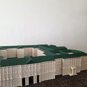 May include: A white architectural model with a green roof, showcasing a detailed building design. The model includes numerous windows, arched entrances, and a symmetrical layout, possibly representing a historical or classical structure. It rests on a textured surface.