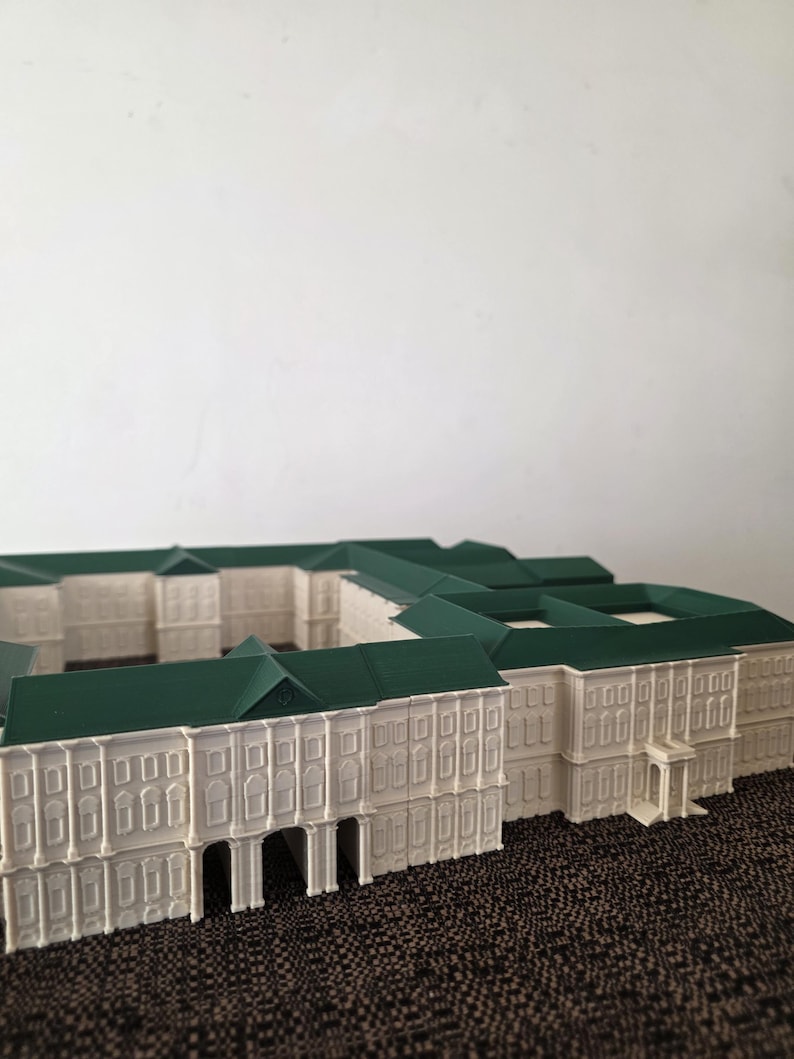 May include: A white architectural model with a green roof, showcasing a detailed building design. The model includes numerous windows, arched entrances, and a symmetrical layout, possibly representing a historical or classical structure. It rests on a textured surface.