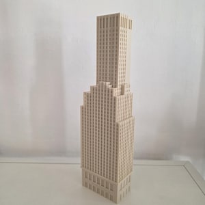 Può includere: Un modello architettonico tridimensionale color beige di un grattacielo con un design a gradoni e una torre centrale che si eleva sopra una base più ampia. La struttura è coperta da una griglia di piccole finestre quadrate incassate.