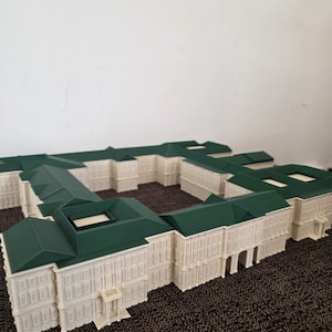May include: A detailed architectural model of a building with a white facade and a green roof. The model has multiple sections, showcasing windows, doors, and intricate design elements. The building is set on a textured surface.