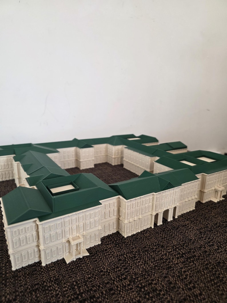 May include: A detailed architectural model of a building with a white facade and a green roof. The model has multiple sections, showcasing windows, doors, and intricate design elements. The building is set on a textured surface.
