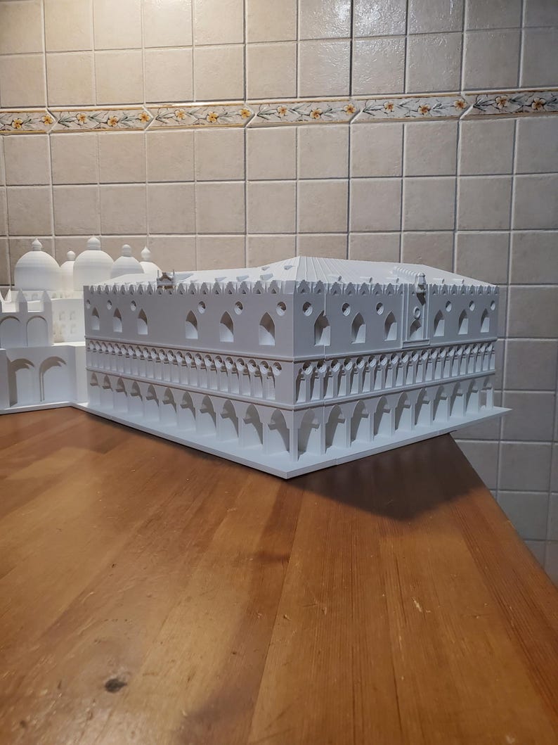 May include: A white 3D printed model of a building with intricate details and arches. The model is sitting on a wooden surface.
