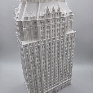 May include: A white 3D-printed model of a tall building, showcasing detailed architectural features. The model has numerous storeys, windows, and a detailed roof. The building's facade is a consistent white, and the model is set against a plain background.