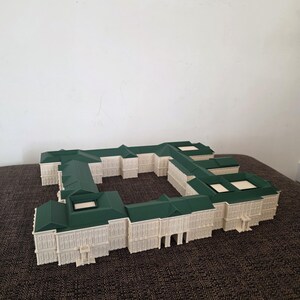 May include: An architectural model of a building with a green roof and off-white walls. The model has a rectangular shape with multiple sections and windows. The building sits on a brown surface.