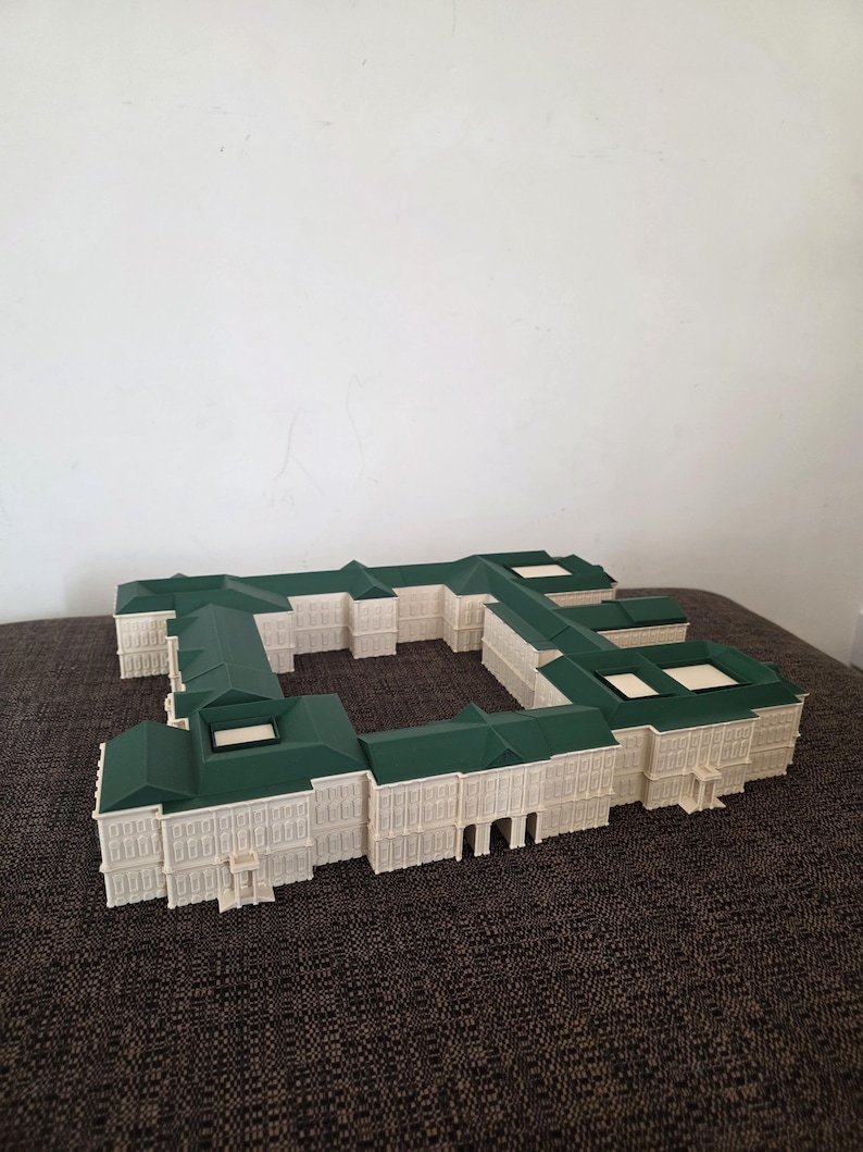 May include: An architectural model of a building with a green roof and off-white walls. The model has a rectangular shape with multiple sections and windows. The building sits on a brown surface.