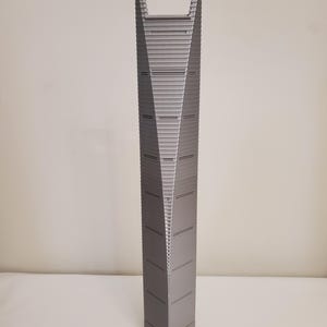 May include: A grey 3D-printed model of a skyscraper. The building features a distinctive, twisting design with horizontal lines and a rectangular opening at the top. The model is a solid, architectural representation.