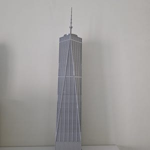 May include: A grey model of the One World Trade Center skyscraper. The building is a tall, rectangular structure with a spire at the top. The facade features a grid-like pattern of windows and vertical supports. The model is set against a plain white background.