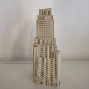 Könnte beinhalten: Ein beiges, dreidimensionales Architekturmodell eines Wolkenkratzers mit abgestuftem, mehrstufigem Design. Die Struktur zeigt ein Raster aus gleichmäßigen, vertieften rechteckigen Fenstern an der Fassade und einen oberen Bereich mit bogenförmigen Öffnungen.