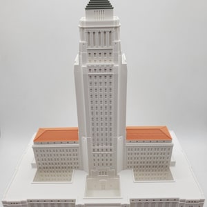 May include: A white 3D printed model of the Los Angeles City Hall building. The model is detailed with a green roof, orange windows, and a white base.
