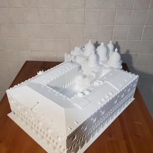 May include: A white 3D printed model of a building with multiple domes on the roof. The model is detailed with intricate architectural features and is displayed on a wooden surface.