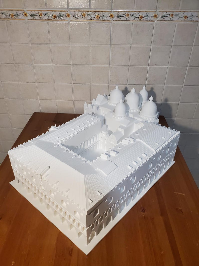 May include: A white 3D printed model of a building with multiple domes on the roof. The model is detailed with intricate architectural features and is displayed on a wooden surface.
