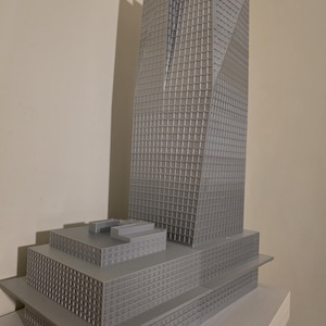May include: A gray 3D printed architectural model of a skyscraper featuring a tall, multi-faceted tower rising from a wide, multi-level rectangular base. The structure displays a grid-like pattern of windows across all exterior surfaces.