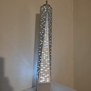 May include: A detailed model of a skyscraper with illuminated windows. The building is tall and rectangular, with a spire at the top. The windows are a light blue colour, and the building is made of a grey material.
