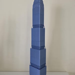 Puede incluir: Maqueta arquitectónica azul de un rascacielos. El modelo es una estructura alta y vertical con detalles intrincados, que incluyen líneas verticales y una parte superior puntiaguda. La base es cuadrada y el diseño general es geométrico.