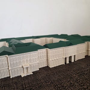 May include: A detailed architectural model of a building with a white facade and green roof. The model features multiple windows and architectural details, showcasing a classical design. The model is placed on a textured brown surface.