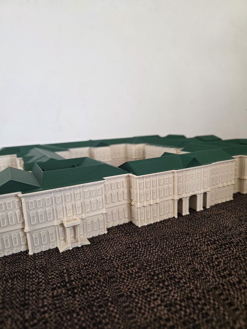 May include: A detailed architectural model of a building with a white facade and green roof. The model features multiple windows and architectural details, showcasing a classical design. The model is placed on a textured brown surface.