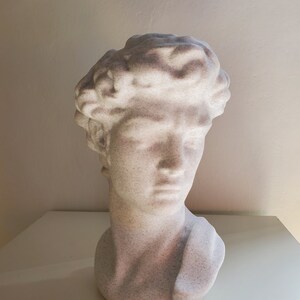 May include: White plaster bust sculpture of a man with a short, curly hairstyle. The bust is a simple, modern design.
