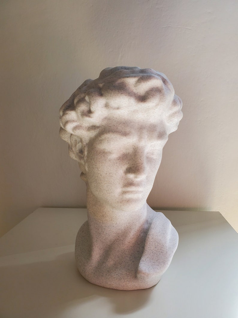 May include: White plaster bust sculpture of a man with a short, curly hairstyle. The bust is a simple, modern design.