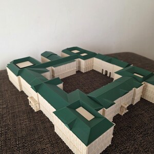 May include: A detailed architectural model of a building with a green roof and off-white walls. The model has a complex design with multiple sections and a courtyard. The roof is a dark green colour, and the walls are a light cream colour.