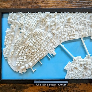 Könnte beinhalten: Ein weißes 3D-gedrucktes Modell von Manhattan, New York City, mit blauem Hintergrund. Das Modell ist in Schwarz gerahmt und enthält den Text "Manhattan NYC".