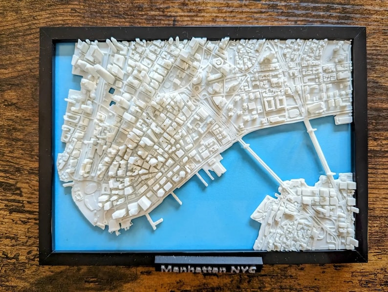 Op de afbeelding: Een wit 3D-geprint model van Manhattan, New York City, met een blauwe achtergrond. Het model is ingelijst in zwart en bevat de tekst "Manhattan NYC".