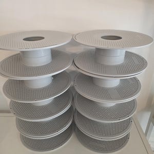 Könnte beinhalten: Zwei vertikale Stapel aus weißen Kunststoff-Filament-Spulen, jede mit einem kreisförmigen Design, einer perforierten Oberfläche und einer zentralen Nabe. Die Spulen sind in zwei Säulen angeordnet und zeigen eine einheitliche Bauweise.