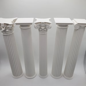 May include: Five white decorative columns with varied capital designs. The columns are fluted and feature different top ornamentation, including floral, square, and scroll patterns. The columns are arranged in a row, showcasing architectural details.