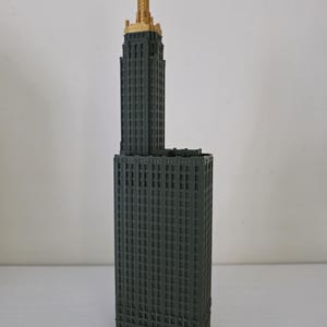 Può includere: Un modello architettonico grigio scuro di un grattacielo con una parte superiore dorata. L'edificio ha una base rettangolare e una torre più alta e stretta. Il modello è dettagliato con molte piccole finestre.