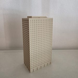 May include: A beige, rectangular skyscraper model featuring a grid of recessed square windows on its exterior walls. The structure has a flat roof with a slightly raised central section and a base with vertical panel detailing on all visible sides.