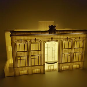 Op de afbeelding: Een gedetailleerd model van een gebouw met een decoratieve gevel, met meerdere ramen en een centrale ingang. Het model is van binnenuit verlicht, waardoor een warme gloed ontstaat.