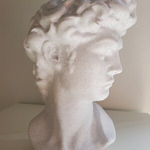 May include: A white plaster bust sculpture of a person with a textured surface. The bust is of a person's head and shoulders, with the person's hair styled in a short, wavy style.